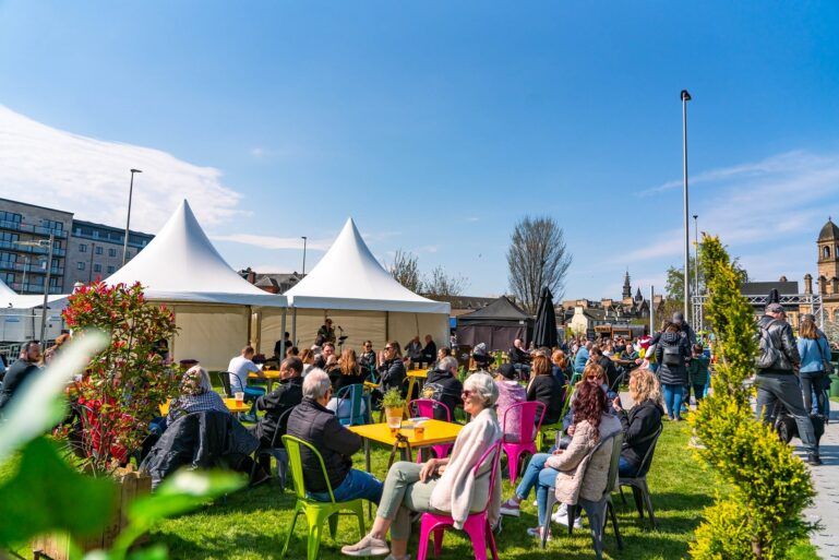 A crowd shot of Paisley Food and Drink Festival 2023 featuring blue skies and people sitting at brightly coloured tables.