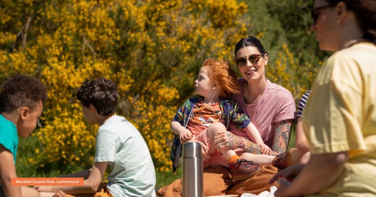 Family enjoy an outdoor picnic outside on a sunny day. A woman wearing sunglasses and pink top is cuddling a young boy.