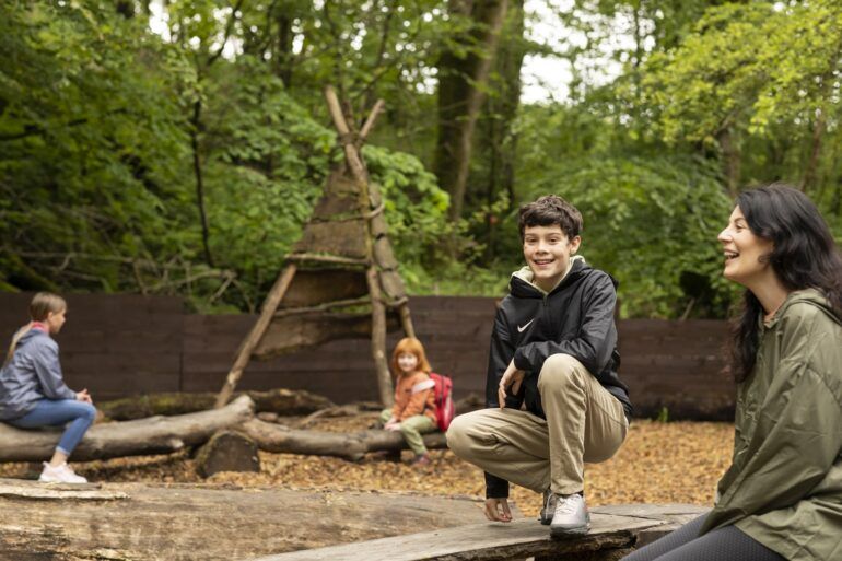 Boy looks to camera while sitting in adventure plaground. Woman and two other children sit alongside