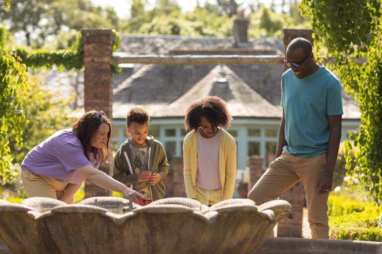 Two adults and two children look at water fountain outside country house