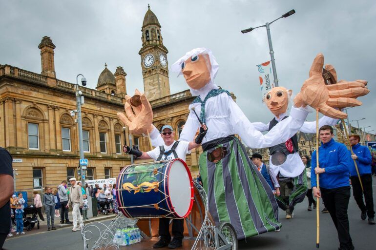 People gather to watch the Sma' Shot Parade travel through Paisley town centre. The parade is led by a man playing a Charleston Drum followed by an oversized puppet dressed as a mill worker. 