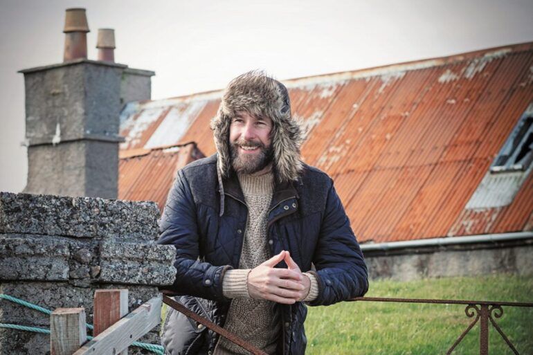 Image shows The Hebridean Baker, Coinneach MacLeod, smiling. He is standing in front of a house with an orange roof