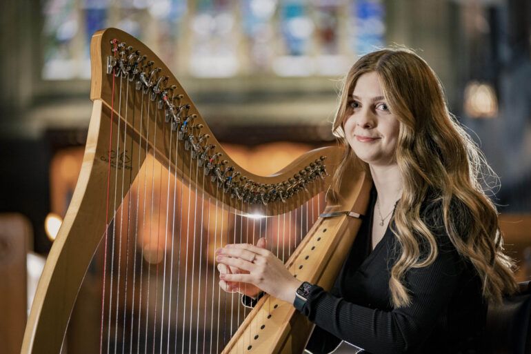 Young girl with long wavy blonde hair is sitting playing a harp while smiling to the camera.