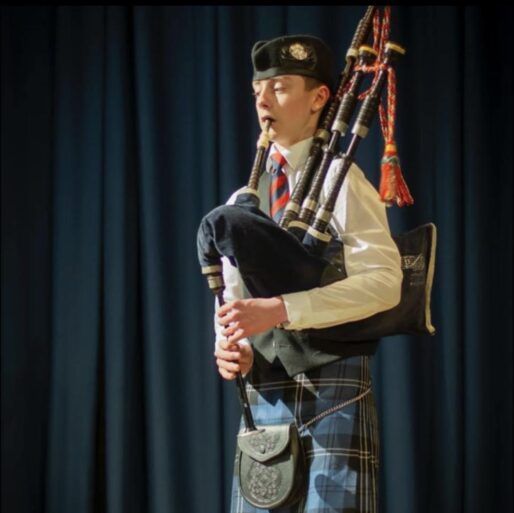 Young male piper aged 16 is dressed in a kilt, shirt, tie and hat and is playing the bagpipes.