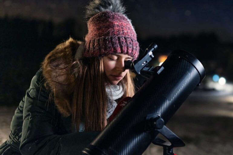 A teenage girl looks through the eyepiece of a telescope pointed up at a dark night sky.
