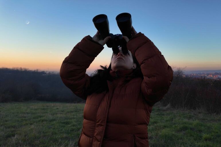 A woman looks at a twilight sky with binoculars.